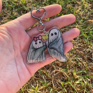 May include: A clear acrylic keyring featuring two cartoon ghosts. One ghost has a pink bow, and both have grey shading. The keyring has a silver heart-shaped clasp. The background is blurred green grass.