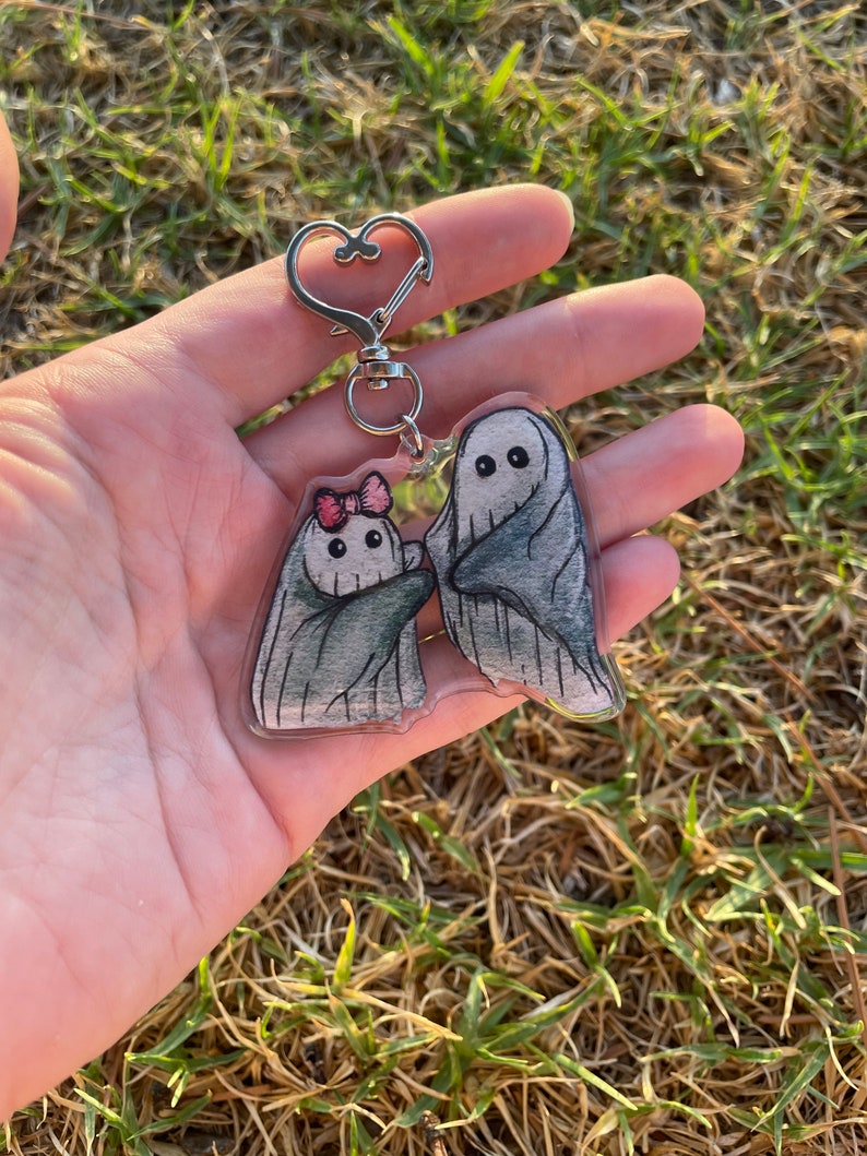 May include: A clear acrylic keyring featuring two cartoon ghosts. One ghost has a pink bow, and both have grey shading. The keyring has a silver heart-shaped clasp. The background is blurred green grass.
