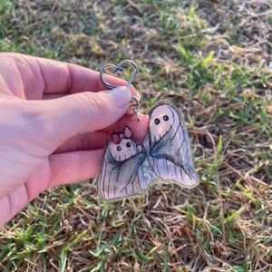May include: A clear acrylic keyring featuring two cartoon ghosts. The larger ghost is wrapped in a grey sheet, and the smaller ghost has a pink bow. The keyring has a silver heart-shaped clasp.