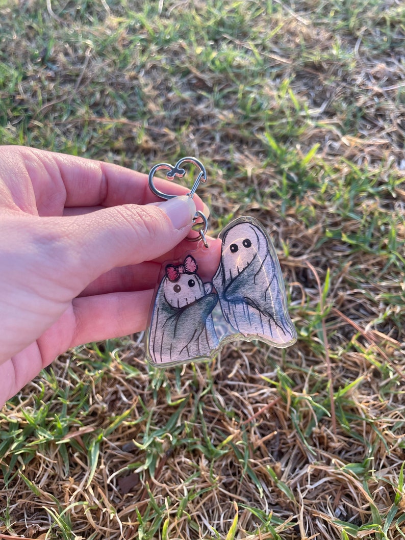 May include: A clear acrylic keyring featuring two cartoon ghosts. The larger ghost is wrapped in a grey sheet, and the smaller ghost has a pink bow. The keyring has a silver heart-shaped clasp.