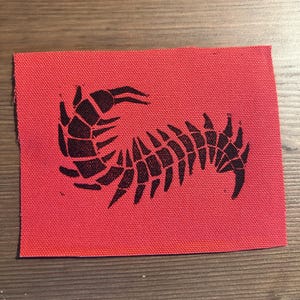 May include: A rectangular fabric patch featuring a vibrant red background with a black, stylized centipede graphic printed in the center. The design displays the segmented body of the insect curving across the surface with multiple legs visible.