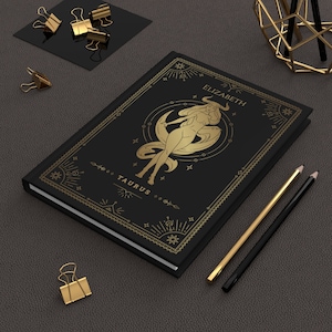 May include: Black notebook with a gold border and a gold illustration of a woman with a star and moon design. The text "TAURUS" and "ELIZABETH" is printed in gold on the cover.