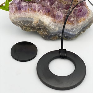 May include: A black obsidian pendant necklace with a round, flat pendant and a smaller, round, flat charm. The necklace is on a black cord and is displayed on a purple amethyst crystal.