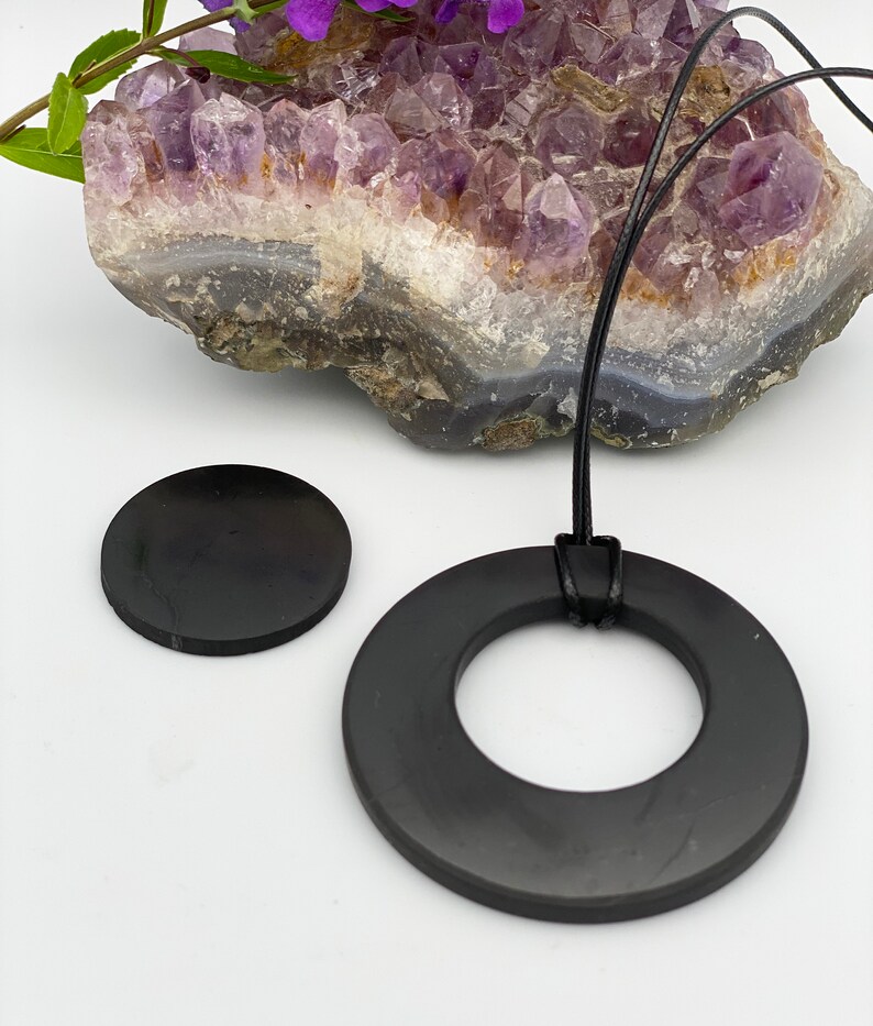 May include: A black obsidian pendant necklace with a round, flat pendant and a smaller, round, flat charm. The necklace is on a black cord and is displayed on a purple amethyst crystal.