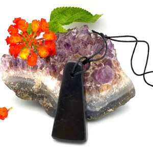 May include: A black pendant with a trapezoid shape hangs from a black cord. The pendant rests on a purple amethyst geode, with orange flowers and green leaves in the background. The pendant is smooth and polished.