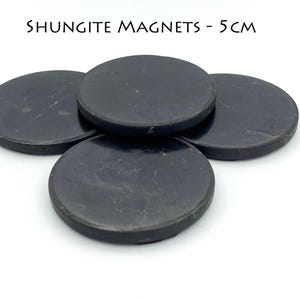 May include: Four round, black shungite magnets, each 5 cm in diameter, are arranged on a white surface. The magnets have a smooth, slightly textured appearance. The text "SHUNGITE MAGNETS - 5 CM" is above the magnets.