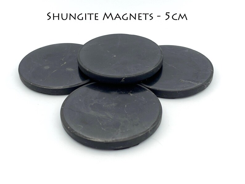 May include: Four round, black shungite magnets, each 5 cm in diameter, are arranged on a white surface. The magnets have a smooth, slightly textured appearance. The text "SHUNGITE MAGNETS - 5 CM" is above the magnets.
