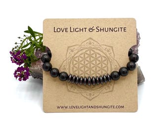 Puede incluir: Pulsera de cuentas negras con un acento de piedra de shungita. La pulsera se muestra en una tarjeta con el texto "Love Light & Shungite" y una dirección de sitio web.