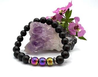 May include: A beaded bracelet with dark brown wooden beads and three iridescent beads. The bracelet is displayed on a purple crystal with pink flowers in the background. The bracelet is a piece of jewelry.