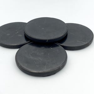 May include: Four black, round coasters with a slightly textured surface. The coasters are made of a dark material and are arranged in a slightly overlapping pattern, showcasing their circular shape and dark color.