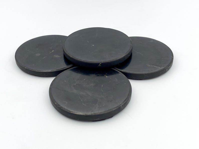 May include: Four black, round coasters with a slightly textured surface. The coasters are made of a dark material and are arranged in a slightly overlapping pattern, showcasing their circular shape and dark color.