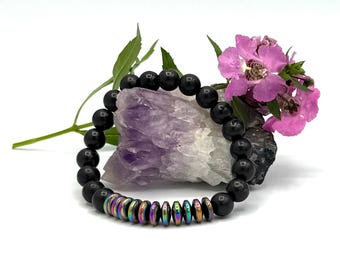 May include: Black beaded bracelet with iridescent hematite beads. The bracelet is resting on a purple amethyst crystal and a green leafy stem with pink flowers.