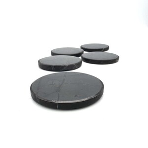 May include: Five dark gray, round coasters are arranged on a white surface. The coasters have a smooth, matte finish and a slightly beveled edge. The coasters are made of a solid, durable material.