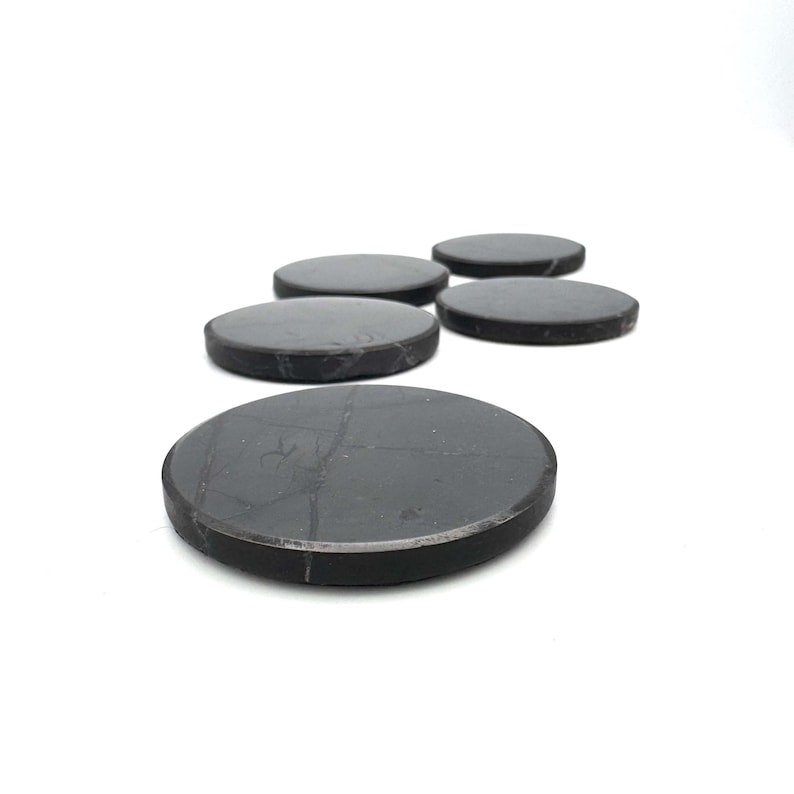 May include: Five dark gray, round coasters are arranged on a white surface. The coasters have a smooth, matte finish and a slightly beveled edge. The coasters are made of a solid, durable material.