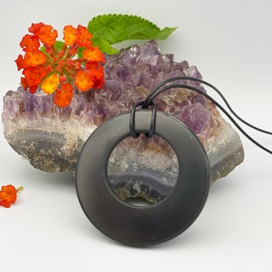 May include: A black obsidian pendant in the shape of a circle with a hole in the center. The pendant is hanging from a black leather cord and is resting on a cluster of amethyst crystals.