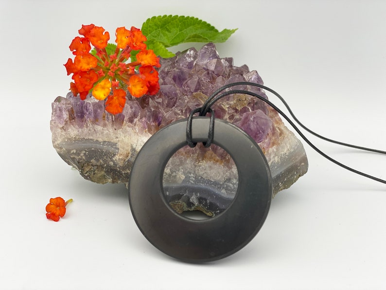 May include: A black obsidian pendant in the shape of a circle with a hole in the center. The pendant is hanging from a black leather cord and is resting on a cluster of amethyst crystals.