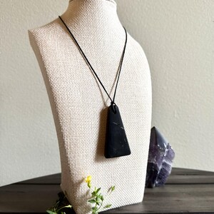 May include: A black pendant necklace with a triangular stone. The stone is suspended from a black cord and displayed on a mannequin. A purple crystal and greenery are in the background.