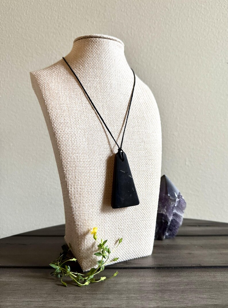 May include: A black pendant necklace with a triangular stone. The stone is suspended from a black cord and displayed on a mannequin. A purple crystal and greenery are in the background.