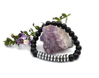May include: Black beaded bracelet with silver-colored metal accents. The bracelet is displayed with a purple crystal and sprigs of greenery and purple flowers. The bracelet is made of round, black beads and silver-colored, flat, circular beads.