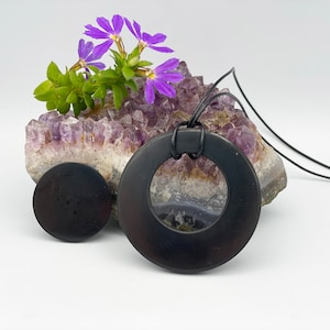 May include: Black obsidian pendant necklace with a round, open circle design. The necklace is displayed on a bed of amethyst crystals with purple flowers.