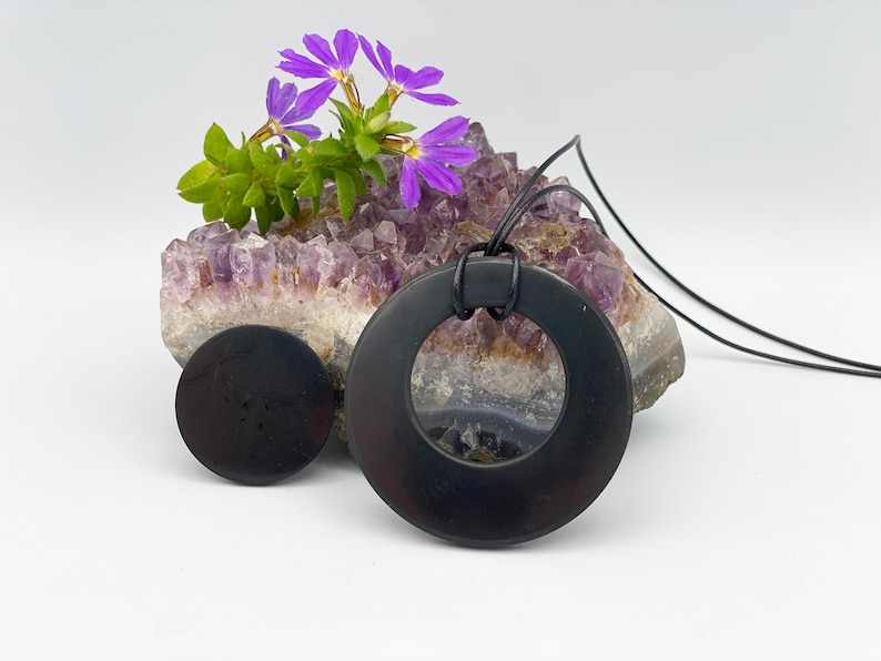 May include: Black obsidian pendant necklace with a round, open circle design. The necklace is displayed on a bed of amethyst crystals with purple flowers.