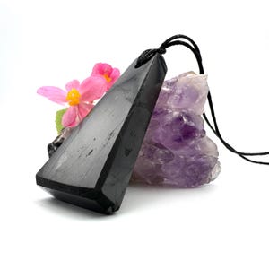 May include: A black, triangular pendant with a black cord, resting against a purple amethyst crystal and pink flowers. The pendant has a polished, reflective surface and a geometric shape, suitable for jewelry.