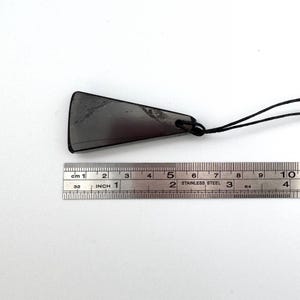 May include: A dark gray, triangular pendant with a black cord. The pendant is approximately 2 inches long. A stainless steel ruler with both inches and centimeters is in the image.