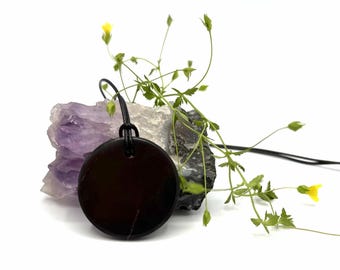 May include: A circular dark brown pendant hangs from a black cord, positioned in front of a jagged, translucent purple amethyst crystal cluster. Delicate green stems with small yellow flowers extend behind the pendant and crystal.