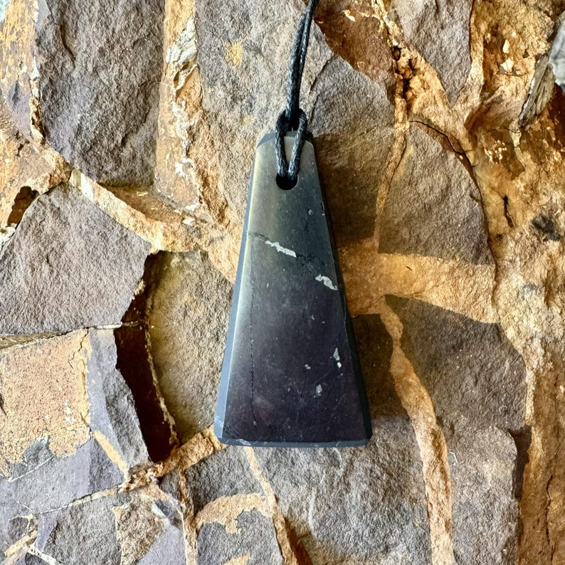 May include: A dark gray, triangular pendant with a black cord. The pendant has a smooth, polished surface with subtle white veining. The pendant is suspended against a textured, brown and gray stone background.