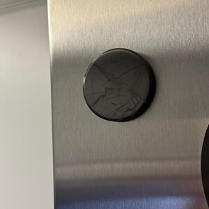 May include: A round, black magnet with a cracked surface is affixed to a brushed metal surface. The magnet is centered on the metal, which has a vertical grain. The background is a plain white wall.