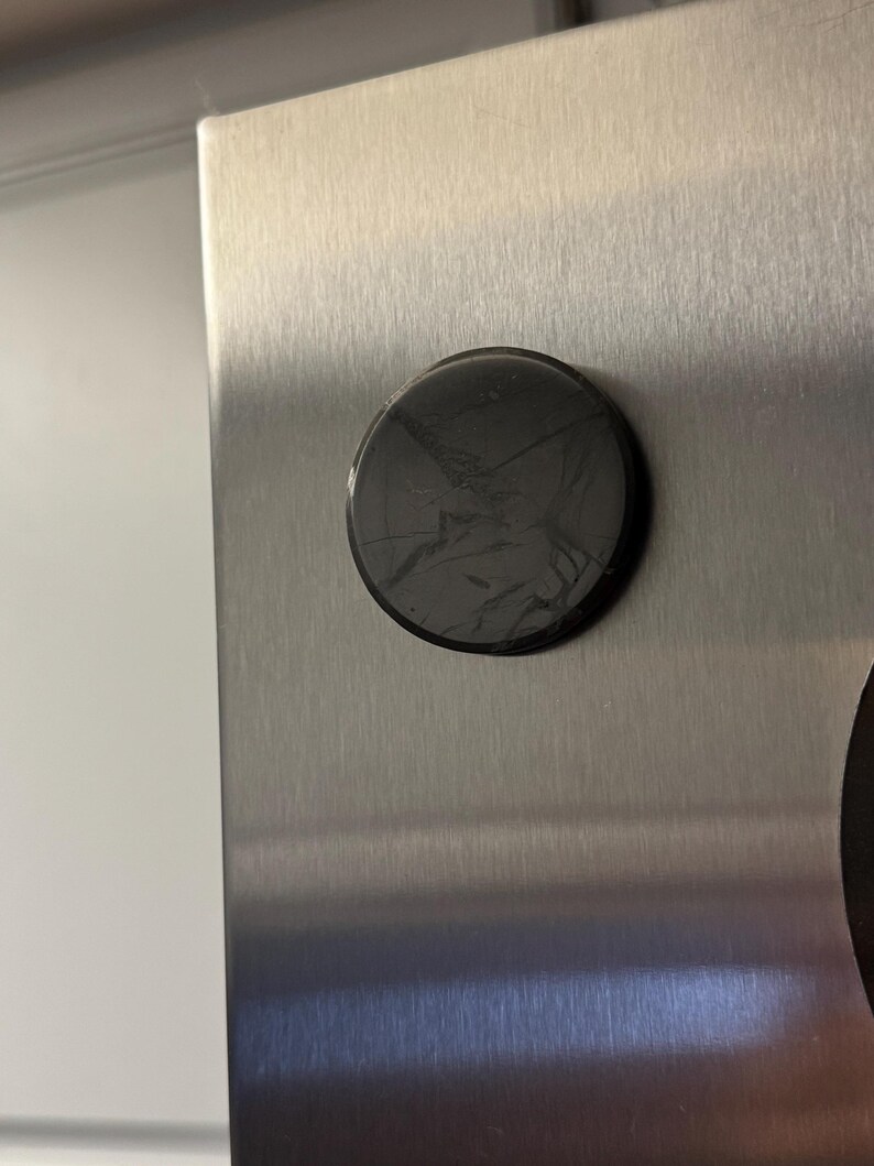 May include: A round, black magnet with a cracked surface is affixed to a brushed metal surface. The magnet is centered on the metal, which has a vertical grain. The background is a plain white wall.