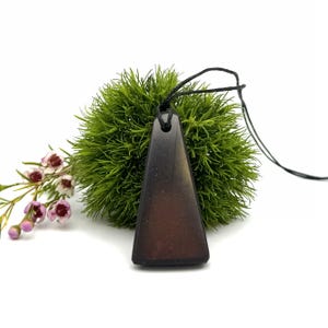 May include: A dark brown triangular pendant necklace with a black cord. The pendant is set against a backdrop of green foliage and small pink and white flowers. The pendant is a solid color with a smooth surface.