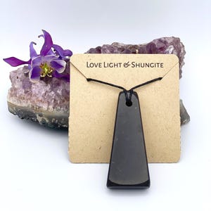 May include: A black, trapezoid-shaped Shungite pendant necklace on a brown card with the text "Love Light & Shungite". The pendant hangs on a black cord. A purple flower and amethyst crystal are in the background.