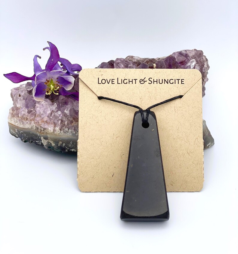 May include: A black, trapezoid-shaped Shungite pendant necklace on a brown card with the text "Love Light & Shungite". The pendant hangs on a black cord. A purple flower and amethyst crystal are in the background.