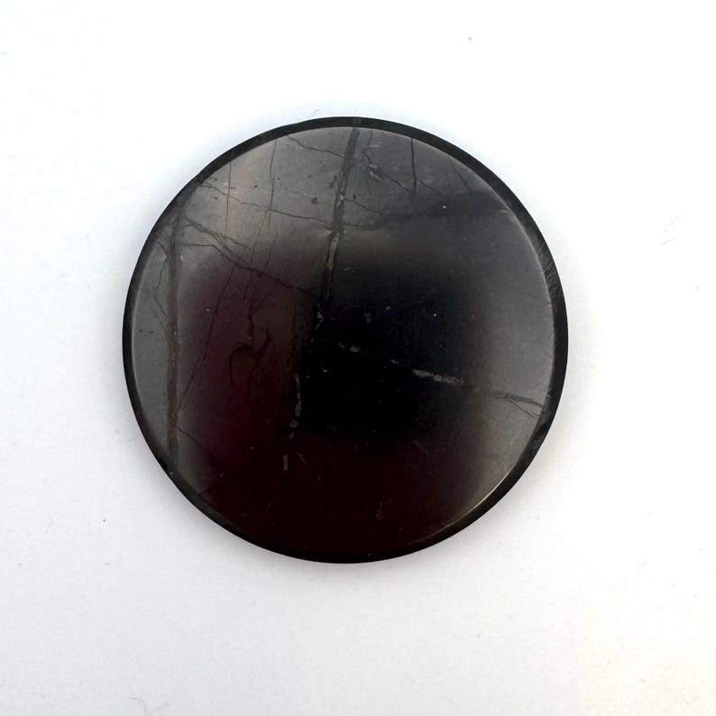 May include: A round, dark gray stone with a polished surface. The stone has subtle, light-colored veining and a smooth, circular shape. The stone is set against a white background.