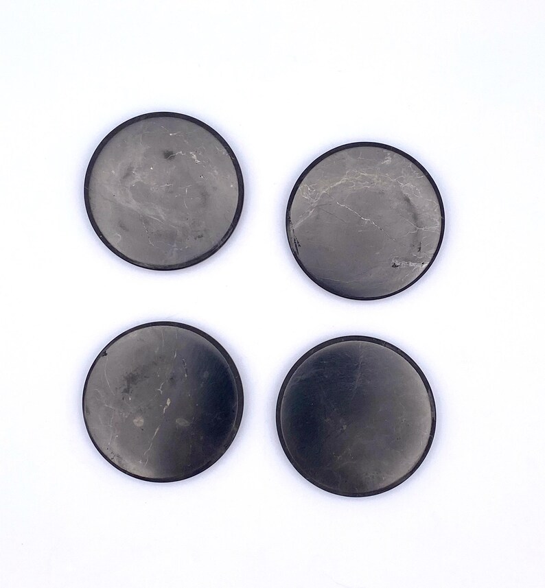 May include: Four round coasters with a dark gray, marbled appearance and a black border. The coasters are arranged in a square formation against a white background. These coasters are designed to protect surfaces from drink rings.