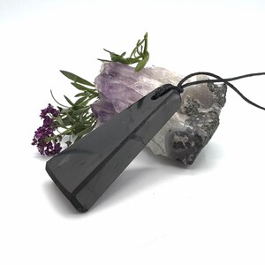 May include: A black, rectangular pendant with a black cord, resting on a white surface. The pendant is angled, showcasing its polished surface. Purple flowers and green sprigs are arranged near the pendant, with a piece of amethyst and a rock in the background.