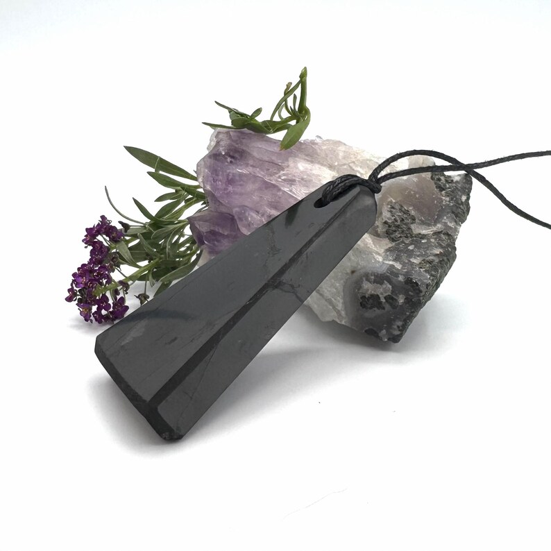 May include: A black, rectangular pendant with a black cord, resting on a white surface. The pendant is angled, showcasing its polished surface. Purple flowers and green sprigs are arranged near the pendant, with a piece of amethyst and a rock in the background.