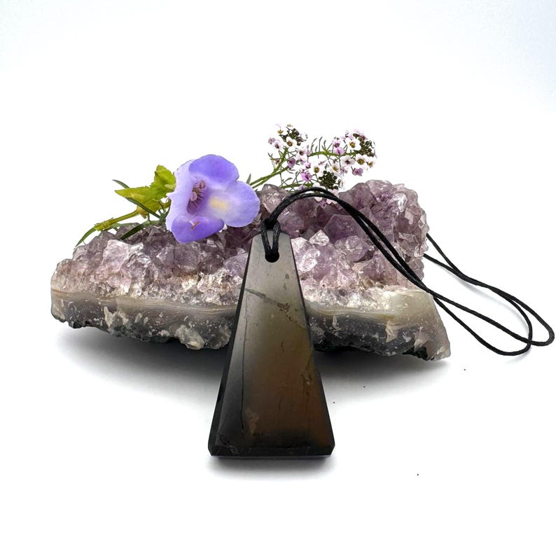 May include: A triangular, dark brown pendant with a black cord hangs in front of a purple amethyst geode. A purple flower and small white flowers are placed on top of the geode. The pendant is made of a smooth, polished stone.