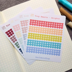 Peut inclure: Un ensemble de cinq feuilles d'autocollants avec des rangées de petits autocollants en forme de cœur de différentes couleurs. Les couleurs sont le rouge, l'orange, le jaune, le bleu et le violet. Les feuilles d'autocollants sont étiquetées "Tiny Hearts - Brights" et "ZeroDesuShop".