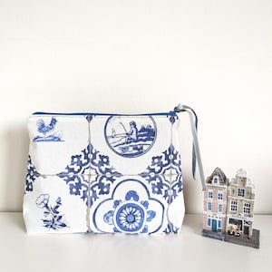 May include: A white zippered pouch with a blue and white Dutch tile pattern. The pouch features a rooster, a fisherman, and floral designs.