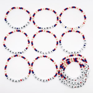 Op de afbeelding: Negen armbanden met kralen met witte letterkralen die namen en cijfers spellen, afgewisseld met rode, blauwe en gouden kralen. De armbanden zijn gerangschikt op een wit oppervlak.