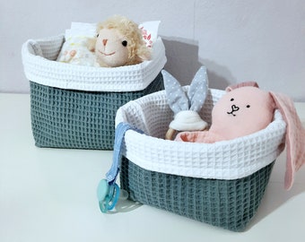Puede incluir: Dos cestas de almacenamiento de tejido de gofre gris y blanco con ribete blanco. Una cesta contiene un cordero de peluche y la otra un conejito de peluche.