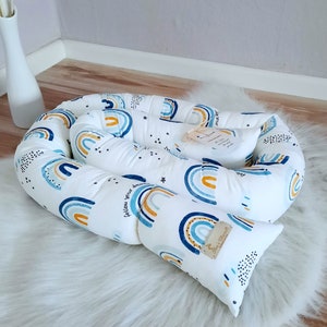 May include: A white baby nest with a blue, orange, and yellow rainbow pattern. The nest includes a matching pillow. The text "follow your dreams" is printed on the side of the nest. The nest is on a white fluffy rug.