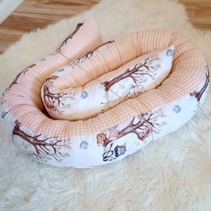 Può includere: Un nido per neonati rosa e bianco con una stampa di animali del bosco. Il nido è realizzato in tessuto morbido e trapuntato ed è perfetto per il comfort e la sicurezza del bambino.