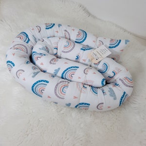 May include: A white, donut-shaped pillow with a rainbow and heart pattern. The pillow is filled with stuffing and is designed for a baby or toddler.