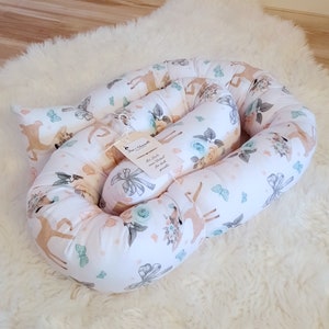 May include: A white and beige baby nest with a deer and floral print. The nest is made of soft fabric and is designed to provide a safe and comfortable sleeping space for a baby. The tag on the nest reads "Mit Liebe zum Detail für dich gewickelt".