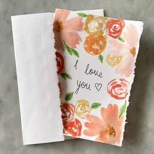 May include: A watercolor greeting card with a floral design in shades of peach, orange, and yellow, with green leaves. The card reads "I love you" in black script. A white envelope is next to the card.
