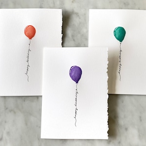 May include: Three handmade birthday cards with watercolor balloon illustrations. Each card features a different colored balloon: red-orange, teal, and purple. The words "happy birthday" are written in cursive below each balloon.