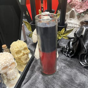 Op de afbeelding: Een zwarte en rode kaars in een glazen pot. De kaars is gelaagd met zwart wax bovenop rood wax. De kaars staat op een grijs, pluche oppervlak.